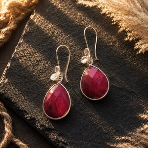 May include: A pair of drop earrings featuring faceted, pear-shaped deep red gemstones set in silver-toned metal. Each earring includes a curved hook wire and a small, delicate silver flower accent positioned above the main teardrop-shaped stone.