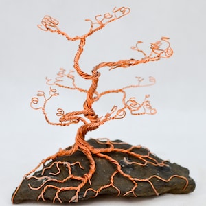 May include: A copper wire tree sculpture, with a twisted trunk and branches with curled ends. The tree is mounted on a dark grey stone base. The copper has a warm, reddish-brown hue, creating a natural, artistic aesthetic. The base is a dark grey stone.