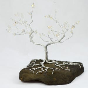 Könnte beinhalten: Eine handgefertigte Drahtbaumskulptur mit einem silberfarbenen Stamm und Ästen. Die Äste sind mit klaren, irisierenden Perlen verziert, die Blüten ähneln. Der Baum ist in einer dunkelbraunen, natürlichen Steinbasis verwurzelt, was eine Miniatur-Bonsai-Ästhetik erzeugt.