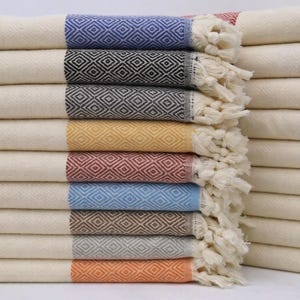 May include: A stack of colorful Turkish towels with diamond patterns and fringed edges. The towels feature stripes of blue, black, gray, yellow, red, light blue, brown, gray, and orange. The towels are made of a soft, absorbent material.
