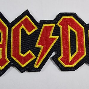 Könnte beinhalten: Ein gestickter Aufnäher in Schwarz, Rot und Gelb mit dem Bandnamen "AC/DC" in einer fetten, stilisierten Schrift.