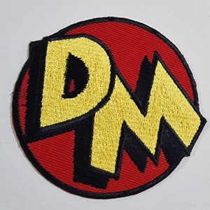 May include: A red circular patch with a black outline. The letters "DM" are embroidered in yellow with black outlines.