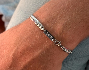 Può includere: Un braccialetto a catena a maglia a catena d'argento con una barra rettangolare con le iniziali "S♡M". Il braccialetto è assicurato con una chiusura a moschettone. Il braccialetto è mostrato su un polso.