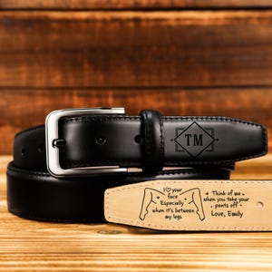May include: A black leather belt with a silver buckle. The belt features a personalized design with the initials "TM" and a romantic message engraved on the inside. The message includes the phrase "I love your face especially when it's between my legs."