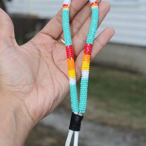 Puede incluir: Lanyard de cuentas hecho a mano en turquesa, rojo, naranja, amarillo y blanco. El lanyard presenta un acento de cuero negro y un llavero plateado. El diseño de cuentas es un accesorio colorido y único.