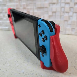 May include: A blue and red Nintendo Switch console with a red 3D printed grip attachment. The console is in handheld mode with the screen facing forward.