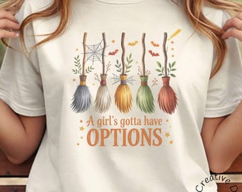 Könnte beinhalten: Cremefarbenes T-Shirt mit fünf bunten Hexenbesen mit dekorativen Elementen wie Spinnweben und Fledermäusen. Der Text "A girl's gotta have OPTIONS" ist in Orange gedruckt. Das Design ist skurril und hat ein Halloween-Thema.