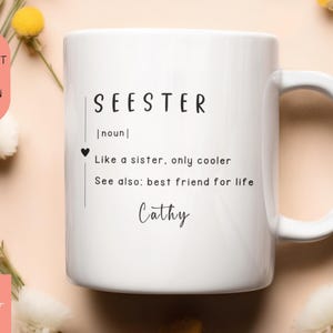 以下が含まれることがあります： 白いセラミック製のマグカップに、黒い文字で「SEESTER [名詞] 姉妹のような存在、でももっとクール。別名：生涯の親友。Cathy」と書いてあります