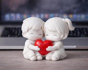 May include: A white figurine of two figures embracing a red heart. The figures are smiling and appear to be a couple. The background includes a laptop computer, suggesting a desk or workspace setting.
