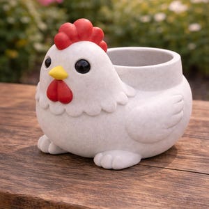 May include: A white ceramic chicken planter with a red comb and wattle, and a yellow beak. The planter has a round body and a large opening for plants. The chicken is sitting on a wooden surface.