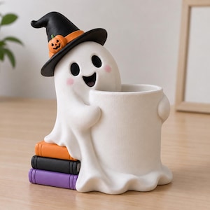 Puede incluir: Una figura de fantasma blanco que lleva un sombrero de bruja negro con un detalle de calabaza naranja. El fantasma tiene mejillas rosadas y sostiene un recipiente cilíndrico blanco mientras está sentado sobre una pila de tres libros naranja, negro y morado.