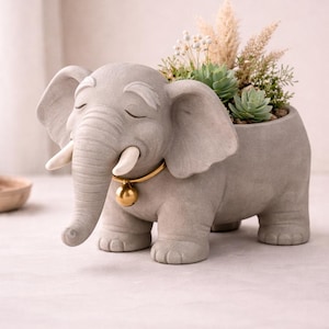 May include: A gray elephant-shaped planter with a gold-colored collar and bell. The elephant has closed eyes and white tusks. The planter is filled with succulents and dried plants. A decorative item for home decor.