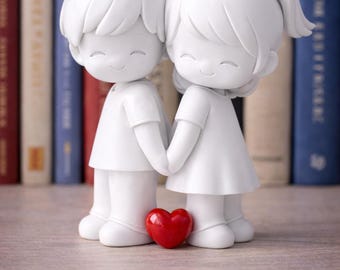 May include: A white figurine of a couple holding hands, with a red heart at their feet. The figures are smiling and dressed in simple clothing. The background features a row of books.