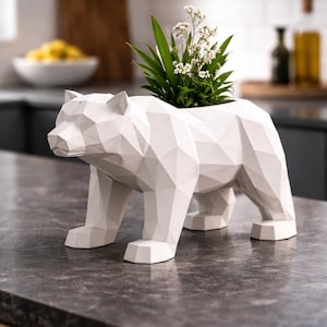 May include: A white geometric bear-shaped planter holding greenery and white flowers. The planter is made of a white material and has a faceted design. The bear is standing on all fours on a dark countertop. The planter is a decorative item for home decor.