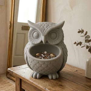 Puede incluir: Un joyero gris con forma de búho hecho de piedra. El búho tiene una gran abertura para guardar joyas, anillos y objetos pequeños. El búho está sobre una mesa de madera. El fondo incluye un espejo y un jarrón con vegetación.
