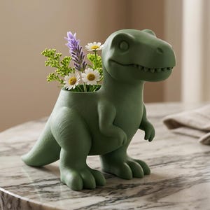 Op de afbeelding: Een groene dinosaurusvormige plantenbak met een klein bloemstuk. De plantenbak is effen, matgroen, met een lachend gezicht en gedetailleerde textuur. De bloemen zijn onder meer witte madeliefjes en paarse lavendel.