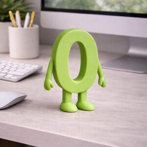 May include: A lime green number zero figurine with arms and legs, standing on a light gray desk. The figurine has a cartoonish design. A computer monitor, keyboard, and mouse are in the background.