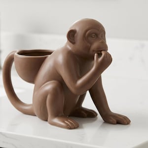 May include: A brown ceramic monkey figurine with a smooth matte finish. The monkey is in a seated position with one hand raised to its mouth. A hollow, bowl-shaped container is attached to the monkey's back, featuring a curved tail handle.