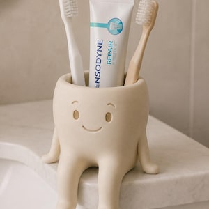 May include: A beige, tooth-shaped toothbrush holder with a smiling face, holding a white toothbrush, a tan toothbrush, and a tube of Sensodyne Repair and Protect toothpaste. The holder features two dangling legs and two small arms on the sides.