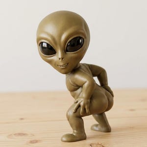 Può includere: Una statuetta aliena color verde oliva opaco con una testa grande, liscia e arrotondata e occhi ovali neri lucidi sovradimensionati. La figura è in posizione accovacciata, inclinata in avanti con una mano sul fianco e un leggero sorriso.