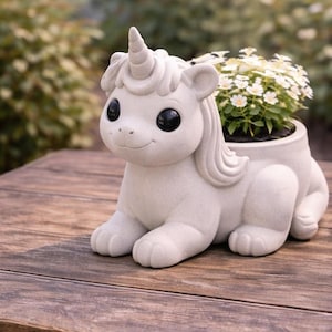 Könnte beinhalten: Ein hellgrauer Einhorn-Blumentopf mit einem lächelnden Gesicht und großen schwarzen Augen. Das Einhorn hat ein spiralförmiges Horn und hält eine kleine Pflanze mit weißen Blüten. Der Topf steht auf einer Holzoberfläche.