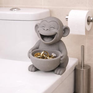 May include: A gray monkey figurine with a bowl-shaped belly, holding several gold rings and a diamond ring. The monkey is smiling widely. The figurine is placed on a white surface, with a toilet and toilet paper roll in the background.