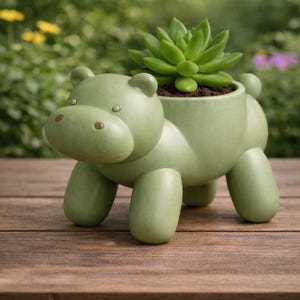 May include: A light green ceramic hippo planter with a succulent plant. The hippo has a rounded body, four stubby legs, and a large head with small ears and eyes. The planter is on a wooden surface.