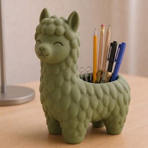 May include: A matte olive green llama-shaped desk organizer with a textured, wool-like surface. The hollow back serves as a container holding several pencils, pens, a metal sharpener, and silver paper clips, all standing upright.