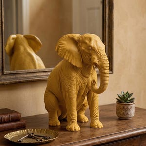 May include: A mustard-yellow elephant figurine with detailed skin texture, seated on a wooden table. The elephant's trunk is curled, and its large ears are visible. A mirror reflects the elephant. A small potted succulent and a decorative tray with keys are also on the table.
