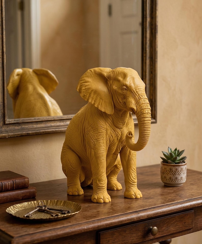 May include: A mustard-yellow elephant figurine with detailed skin texture, seated on a wooden table. The elephant's trunk is curled, and its large ears are visible. A mirror reflects the elephant. A small potted succulent and a decorative tray with keys are also on the table.