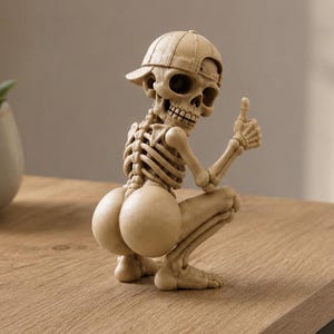 May include: A beige-colored skeleton figurine in a crouching pose, wearing a baseball cap turned backward. The skeleton has a prominent gluteal area and is holding one hand up with the thumb extended in a gesture. The surface is matte and textured.