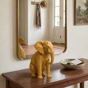 May include: A mustard-yellow elephant figurine sits on a dark wood table. The elephant is detailed with textured skin and large ears. A gold-framed mirror reflects the elephant and the room's interior. A decorative bowl with keys and a card is also on the table.