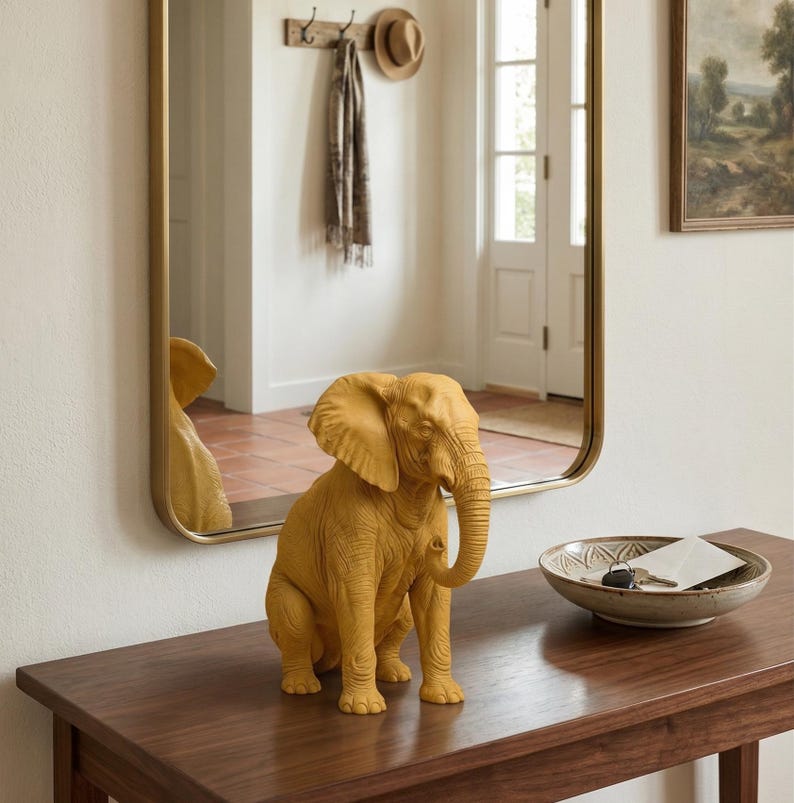 May include: A mustard-yellow elephant figurine sits on a dark wood table. The elephant is detailed with textured skin and large ears. A gold-framed mirror reflects the elephant and the room's interior. A decorative bowl with keys and a card is also on the table.