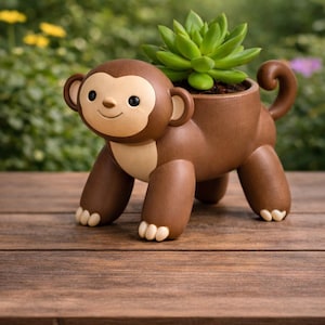 Op de afbeelding: Een bruine apenvormige plantenbak met een lichtgroene vetplant. De keramische plantenbak heeft een lachend gezicht. De aap staat op handen en voeten, met zijn staart opgerold. De plantenbak staat op een houten oppervlak.
