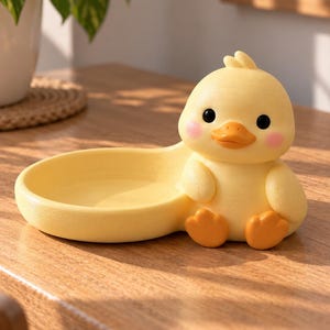 May include: A yellow duck-shaped trinket tray featuring a cartoon-style duck with black eyes, an orange beak, and pink cheeks. The duck is attached to a smooth, shallow, circular yellow dish designed for holding small items or accessories.