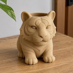 May include: A matte beige tiger-shaped planter with a textured surface. The vessel features a sculpted tiger head with detailed facial markings, pointed ears, and a wide opening at the top for plants. The design shows the tiger in a seated position.