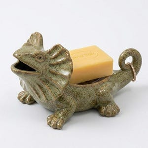 Peut inclure: Un porte-savon en céramique verte en forme de lézard à collerette avec une finition texturée et tachetée. Le lézard a la gueule ouverte et une queue enroulée. Un savon rectangulaire jaune clair Citrus Soap est posé dedans, avec une bague dorée.