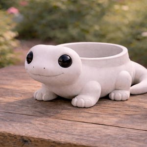 Puede incluir: Una maceta de cerámica blanca con forma de lagarto sonriente con grandes ojos negros. La maceta tiene una abertura redonda para plantas y está sobre una superficie de madera. El fondo está borroso con vegetación.