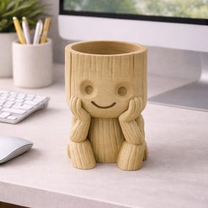 Może przedstawiać: Jasnobrązowa, drewniana doniczka w kształcie postaci z uśmiechniętą twarzą, trzymającą podbródek rękami. Doniczka ma cylindryczny otwór na rośliny i stoi na biurku z komputerem i klawiaturą w tle.