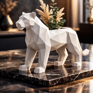 Könnte beinhalten: Ein weißer, geometrischer Löwen-Pflanztopf mit Grünzeug. Die Skulptur besteht aus weißem Material und hat eine ausgehöhlte Rückseite für Pflanzen. Sie steht auf einer dunklen Marmoroberfläche und bildet ein modernes Dekorationsobjekt.
