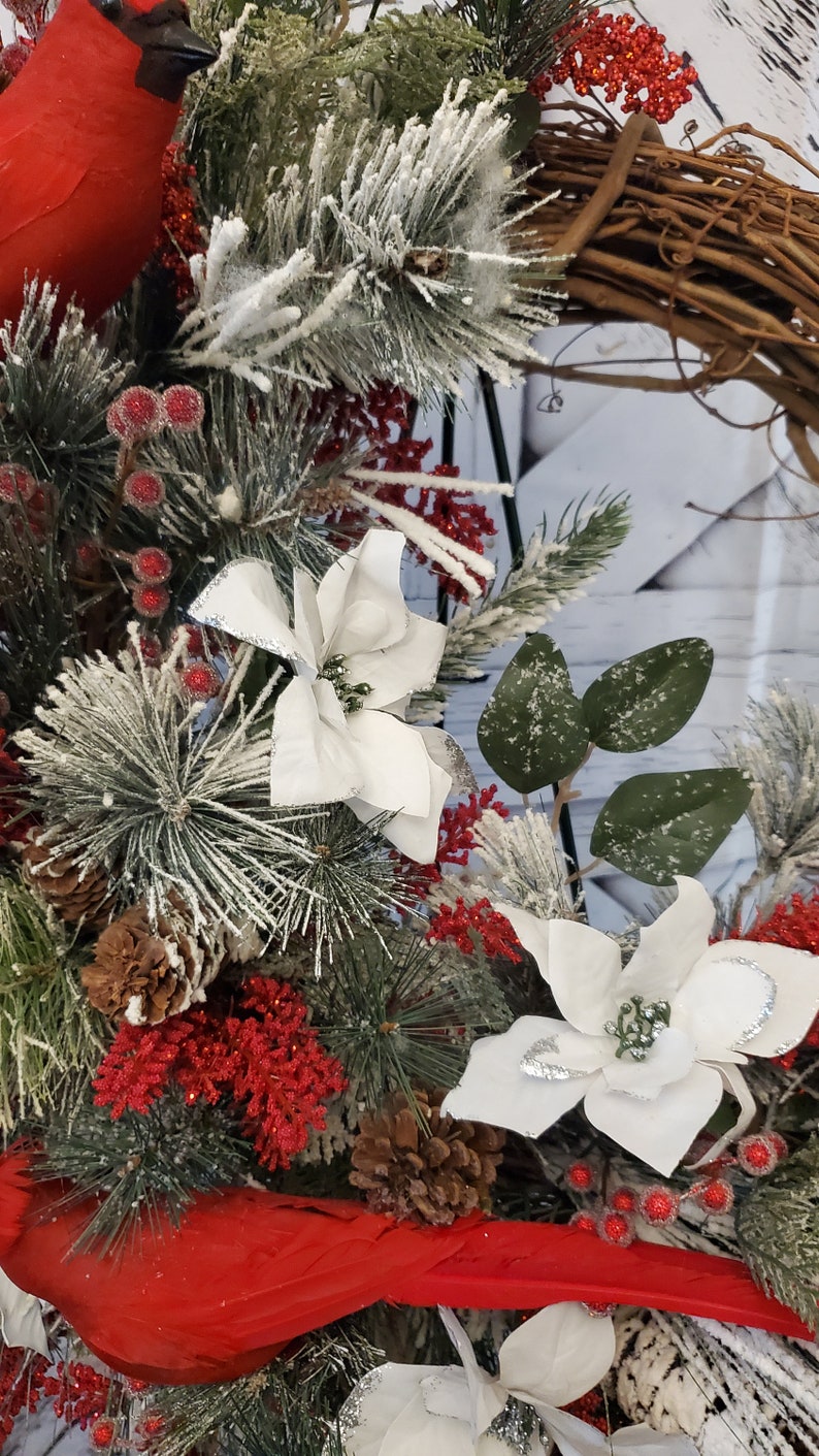 May include: A close-up of a Christmas wreath with a variety of festive elements. The wreath features red cardinals, white poinsettias, pinecones, and red berries. The greenery is dusted with artificial snow, creating a wintery scene.