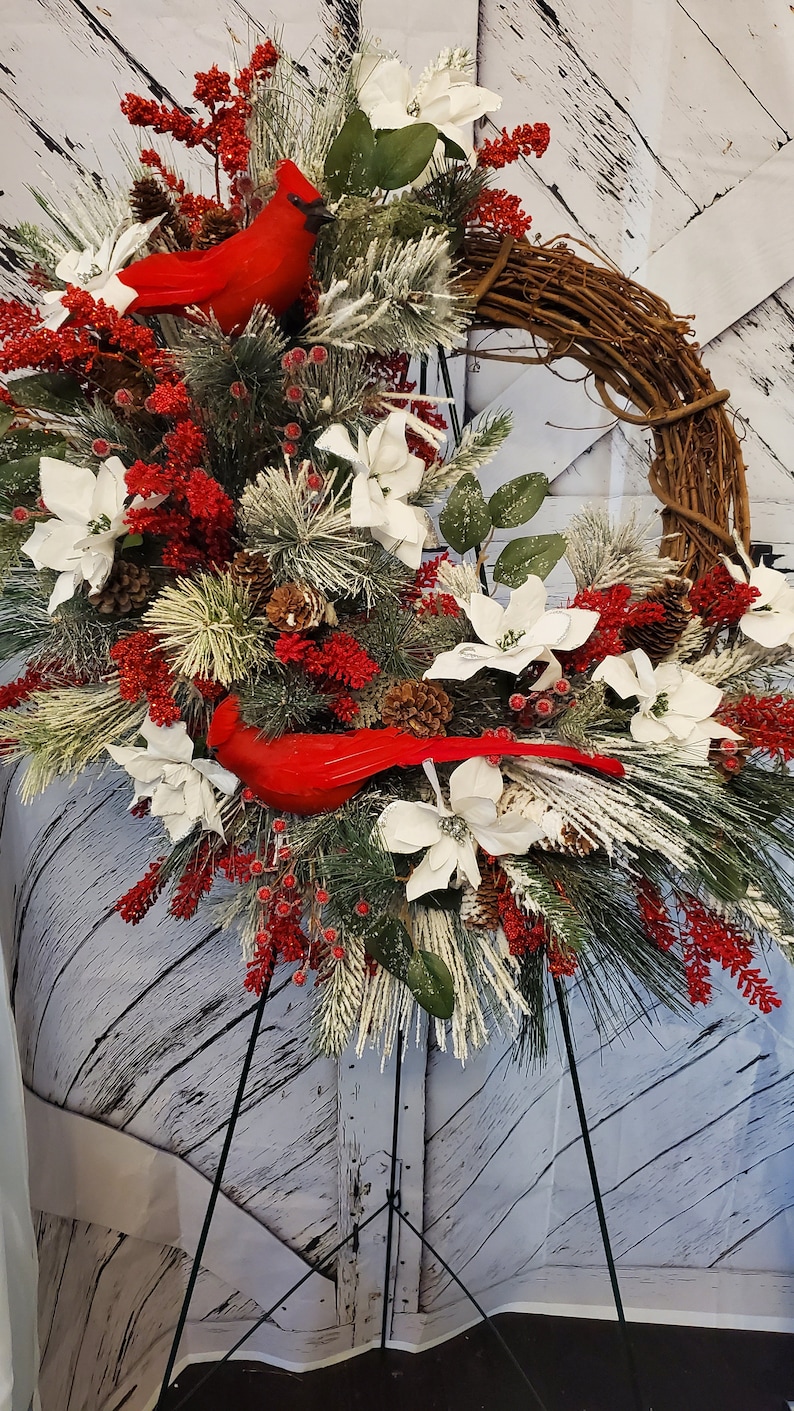 May include: A decorative wreath featuring a grapevine base, adorned with red cardinals, white poinsettias, pinecones, and red berries. The arrangement includes frosted pine branches and is set against a white wooden backdrop.