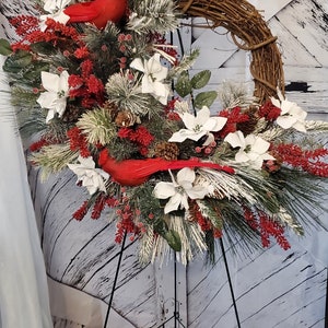 May include: A decorative wreath featuring a grapevine base, adorned with white poinsettias, red berries, and evergreen branches. Two bright red cardinal bird figurines are nestled among the foliage, creating a festive holiday display.