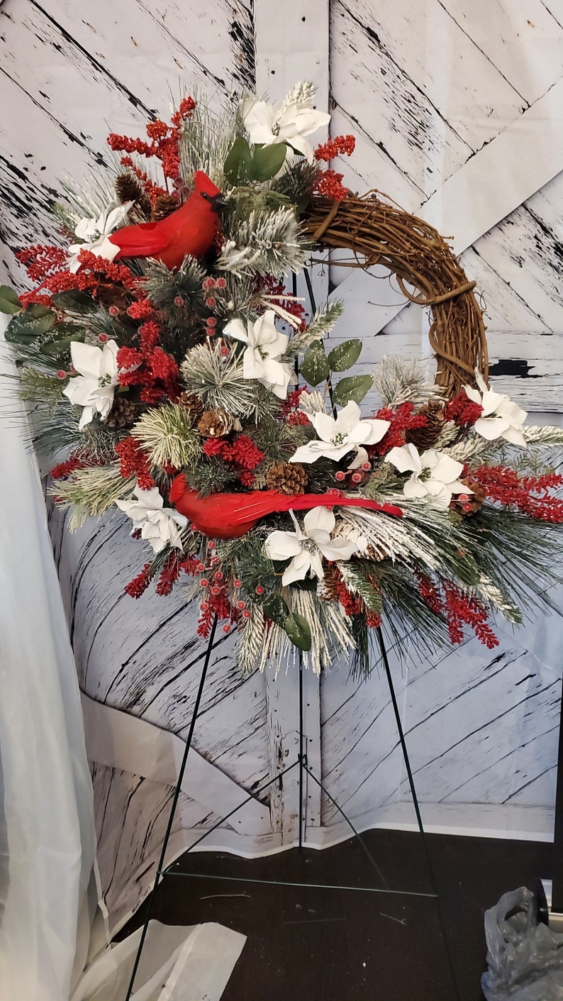 May include: A decorative wreath featuring a grapevine base, adorned with white poinsettias, red berries, and evergreen branches. Two bright red cardinal bird figurines are nestled among the foliage, creating a festive holiday display.