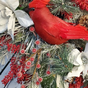 May include: A festive holiday arrangement featuring a vibrant red cardinal bird nestled among frosted pine branches, white poinsettias, and red berry clusters. The arrangement includes pinecones and is set against a light wood background.