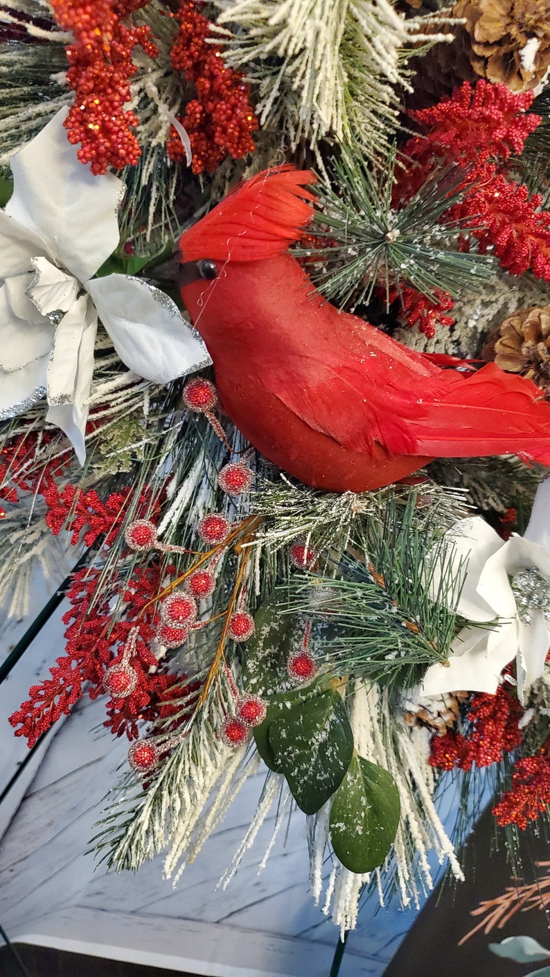 May include: A festive holiday arrangement featuring a vibrant red cardinal bird nestled among frosted pine branches, white poinsettias, and red berry clusters. The arrangement includes pinecones and is set against a light wood background.