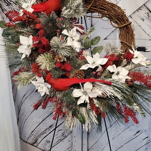 May include: A decorative winter wreath featuring a grapevine base, adorned with white poinsettias, red berries, pinecones, and evergreen branches. Two bright red cardinal bird figurines are nestled within the arrangement. The wreath is displayed on a black metal stand.
