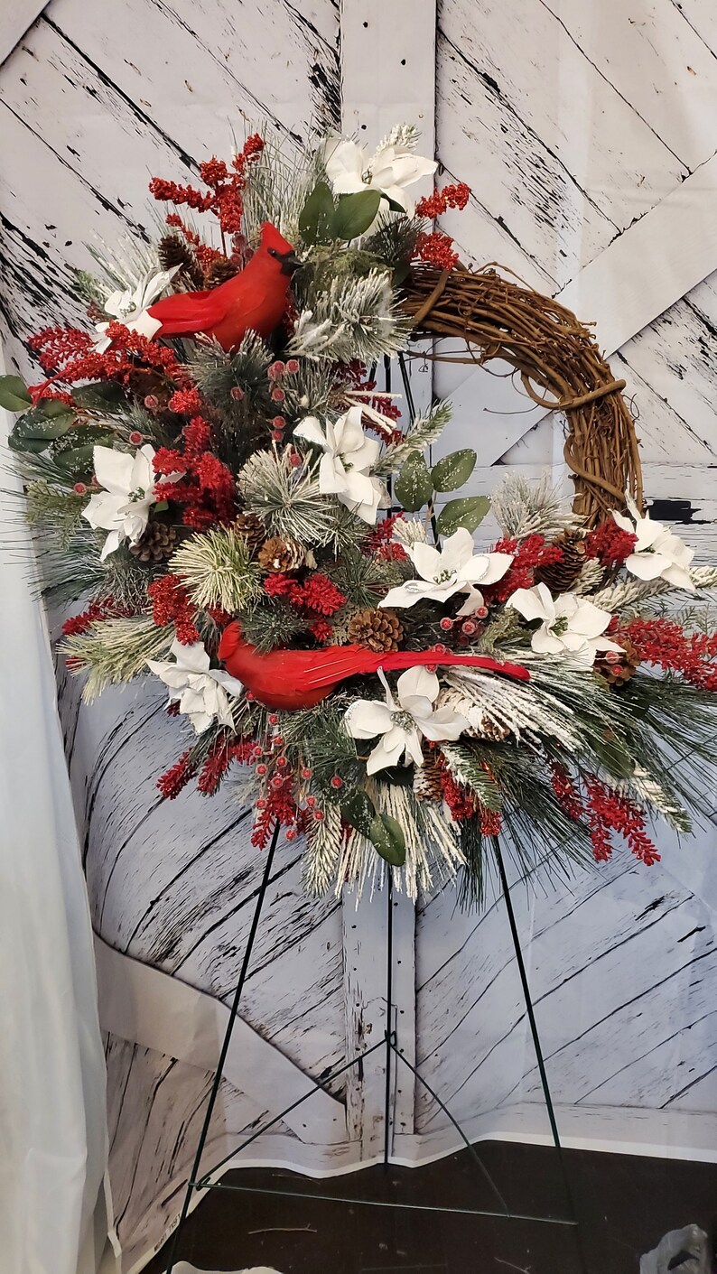 May include: A decorative winter wreath featuring a grapevine base, adorned with white poinsettias, red berries, pinecones, and evergreen branches. Two bright red cardinal bird figurines are nestled within the arrangement. The wreath is displayed on a black metal stand.