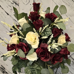 May include: A bouquet of burgundy and ivory roses in a clear glass vase. The roses are arranged in a circular shape and are surrounded by greenery.