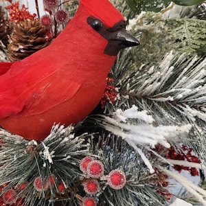 May include: A vibrant red cardinal bird ornament perched amidst frosted evergreen branches, red berries, and pinecones. The bird has a black beak and eye, and is surrounded by white poinsettia flowers. A festive holiday decoration.